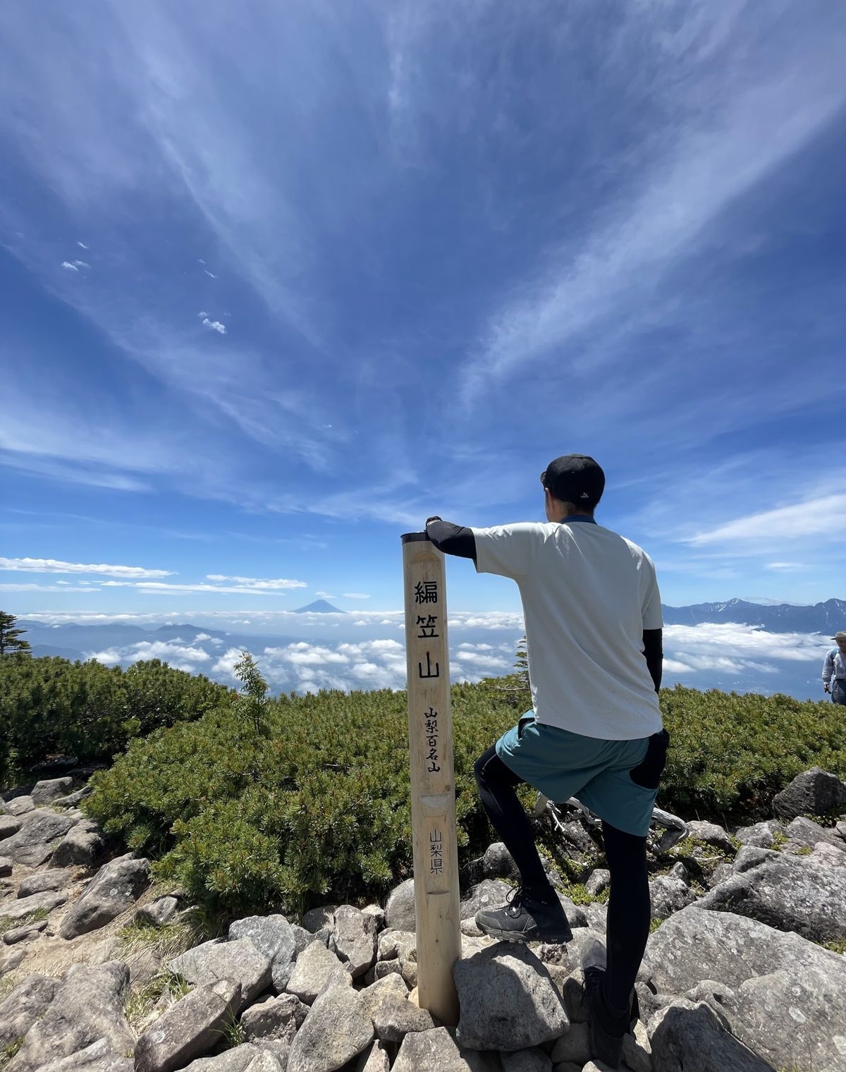 夏の編笠山の山頂標識の横に立つ後ろ姿と、雲海の向こうの富士山
